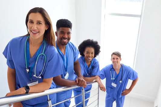 A diverse group of four medical professionals in blue scrubs stand on a staircase, smiling at the camera. They each wear stethoscopes, suggesting their roles as healthcare workers. The background is bright and minimalistic.