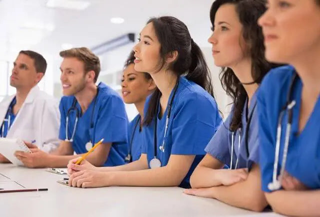 A group of medical professionals, including men and women, sit attentively in blue scrubs with stethoscopes around their necks. They are engaged in a discussion or training session in a bright, well-lit room.