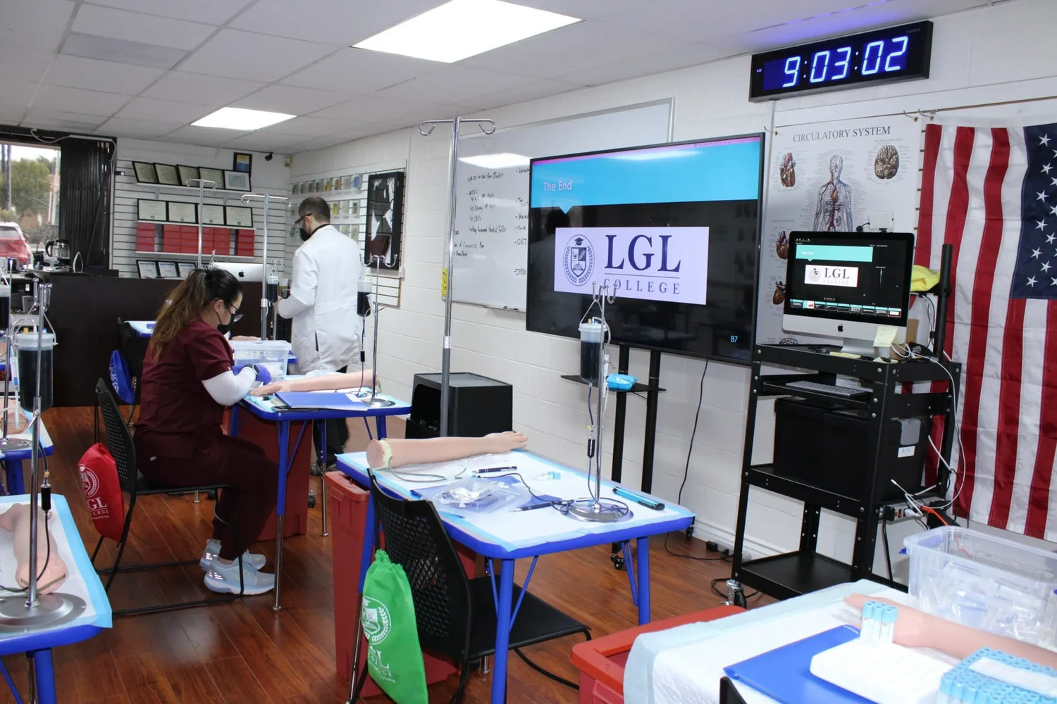 A classroom setup with tables and chairs, featuring medical training equipment, including IV stands and practice arms. Two people in lab coats are engaged in activities. A screen displays