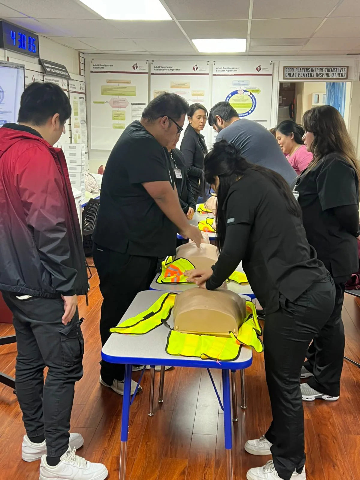 A group of people practicing CPR on mannequins in a classroom setting. They are wearing casual clothing and appear focused on learning the technique. Posters and safety instructions are visible on the walls.