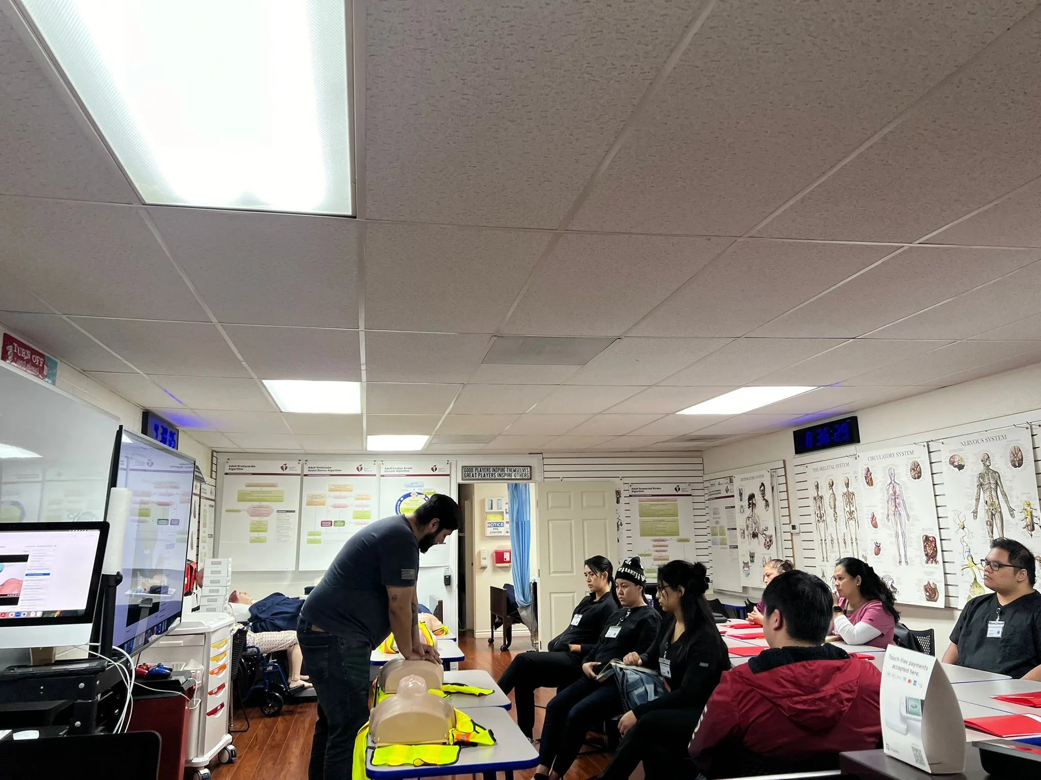 A group of people in a classroom attentively watch an instructor demonstrating a medical procedure on a mannequin. Various medical charts and illustrations are visible on the walls.