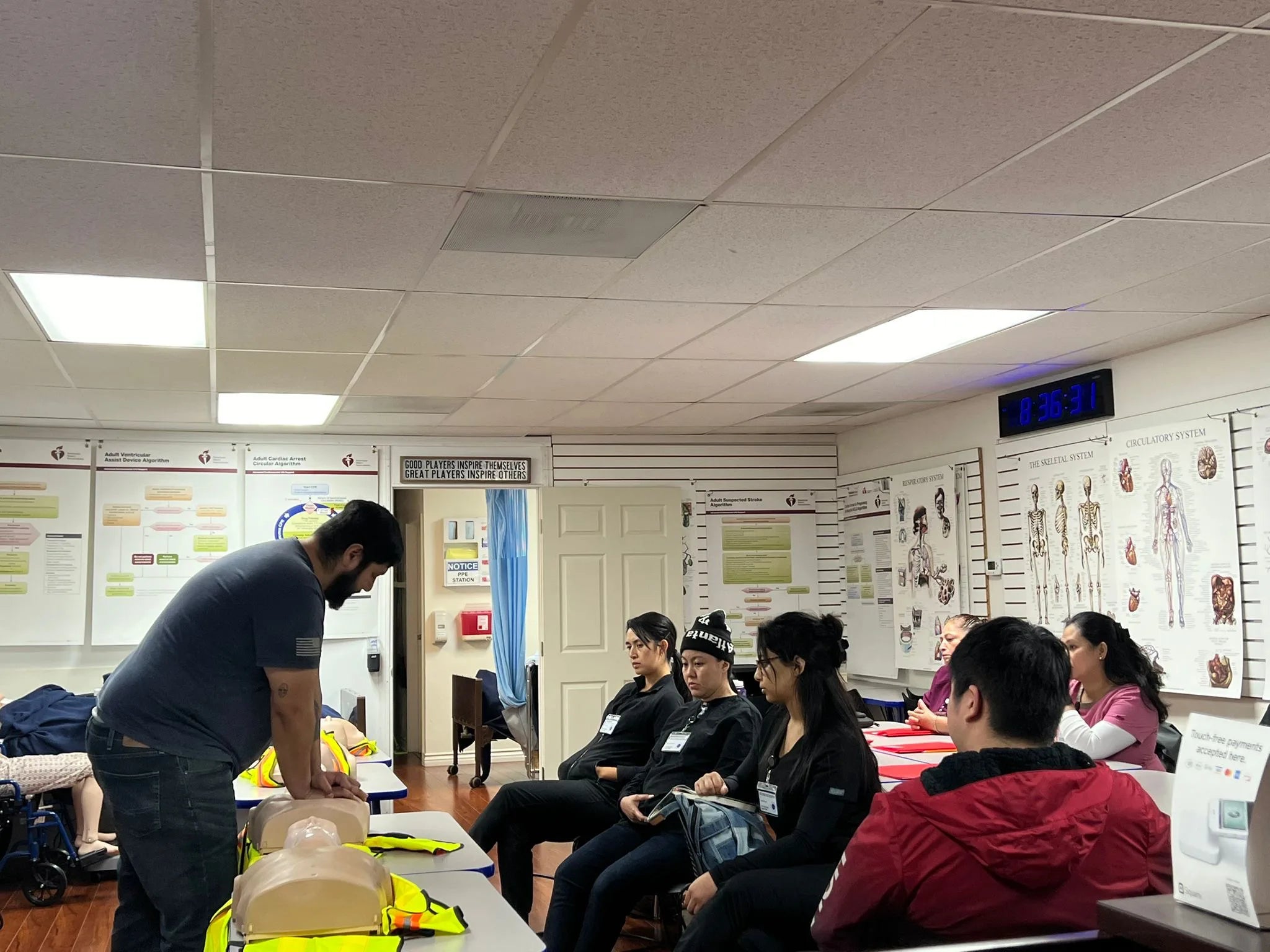 A group of people are sitting in a classroom, attentively watching an instructor demonstrating CPR on a mannequin. The room has educational posters on the walls and a digital clock displaying the time.