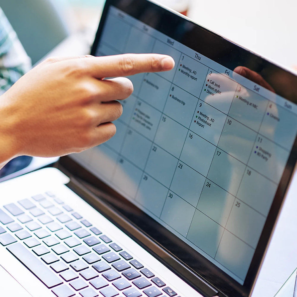 A person points at a laptop screen displaying a monthly calendar with events and appointments listed. The screen shows days of the week from Sunday to Saturday, and the focus is on the middle of the week.